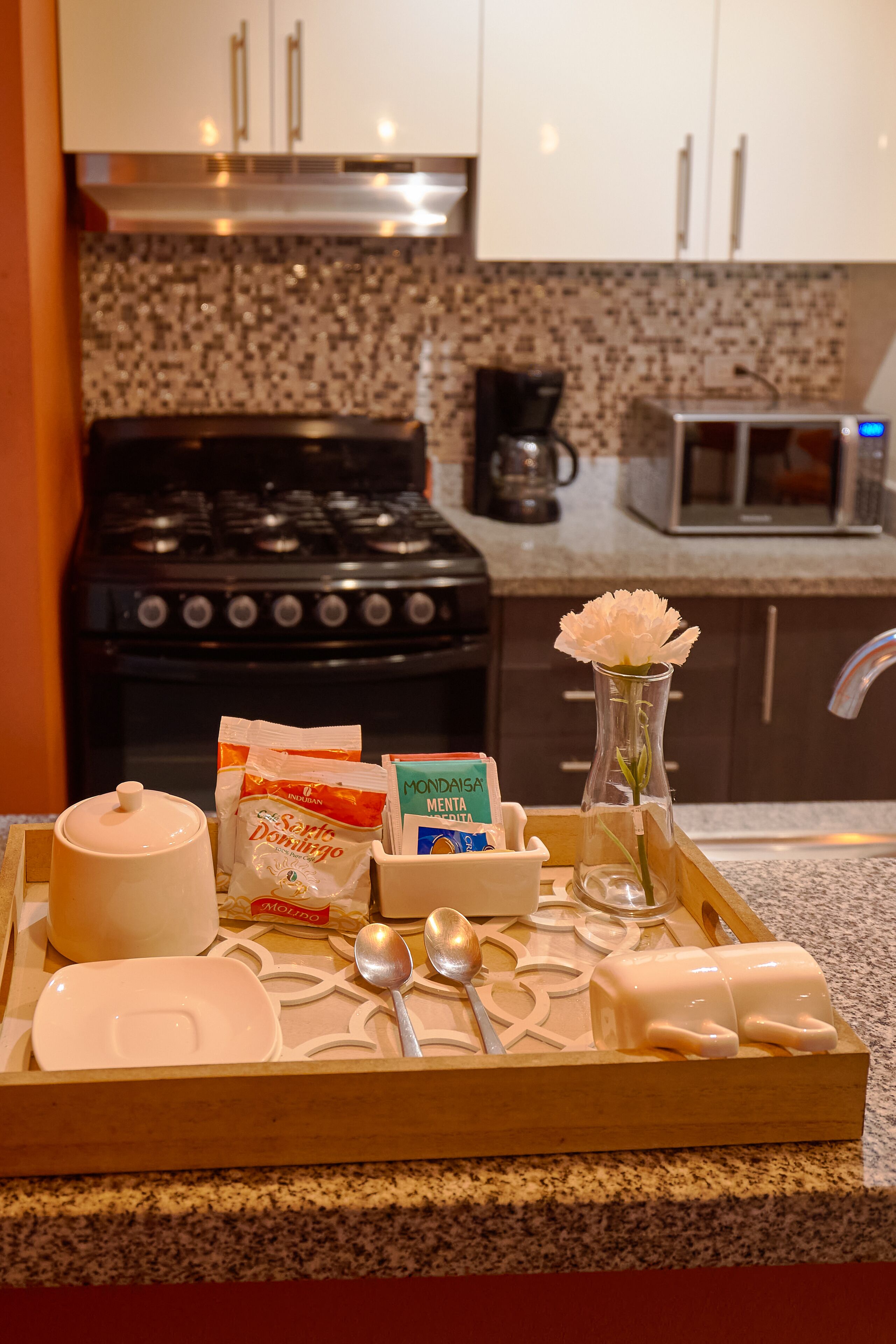 A kitchen counter with a tray containing a white teapot, a vase with a white flower, and various food items.