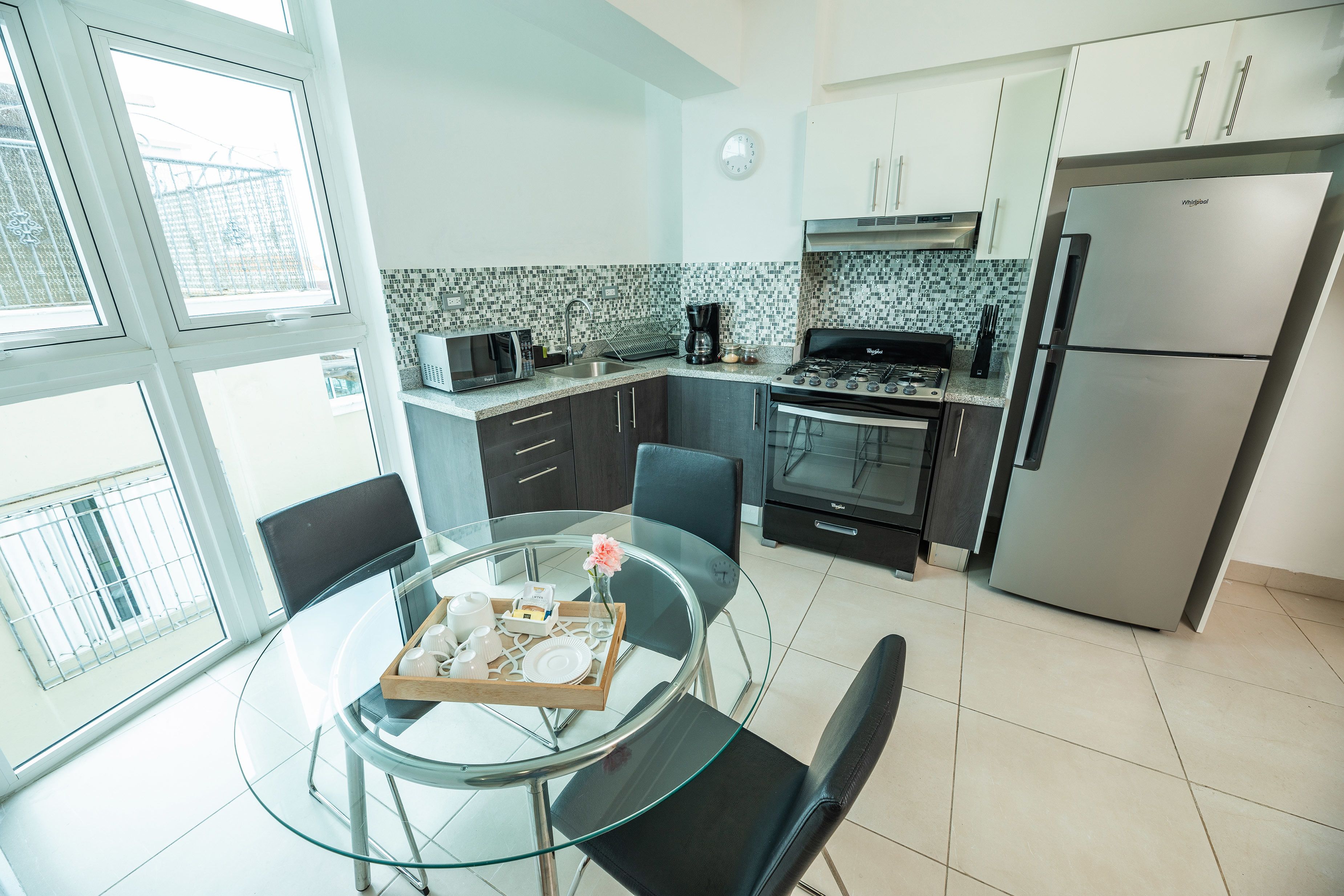 A modern kitchen with a dining area, featuring a glass table, black chairs, a stainless steel refrigerator, and a gas stove.