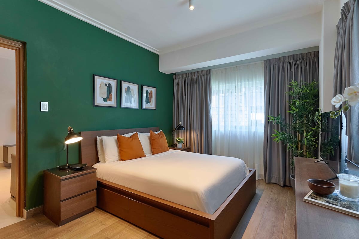 Een moderne slaapkamer met een groot bed, een nachtkastje met een lamp, een bijzettafeltje met een plant en een raam met gordijnen.