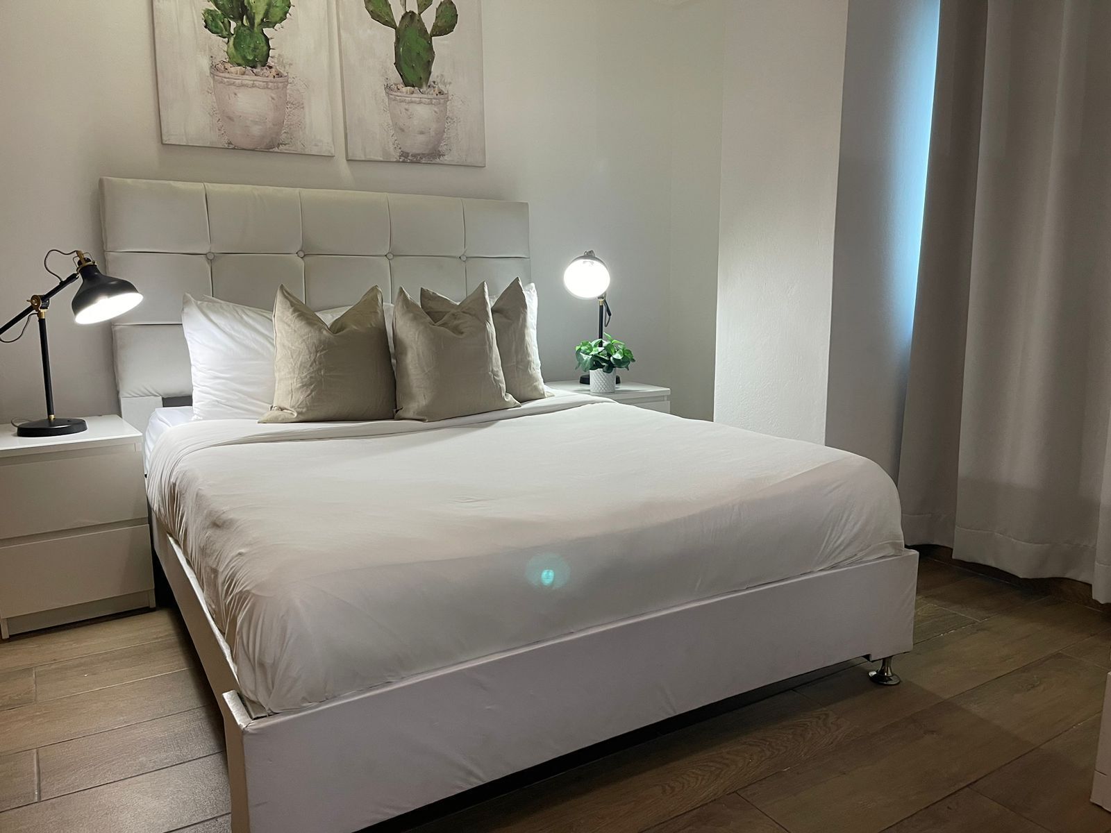 Una cama bien hecha con ropa blanca, cabecera con diseño capitonado, dos lámparas de noche y una pequeña planta en maceta sobre la mesita.