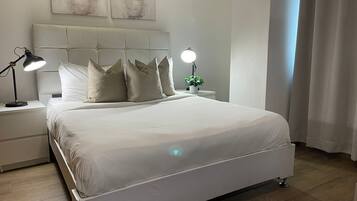 Una cama bien hecha con ropa blanca, cabecera con diseño capitonado, dos lámparas de noche y una pequeña planta en maceta sobre la mesita.