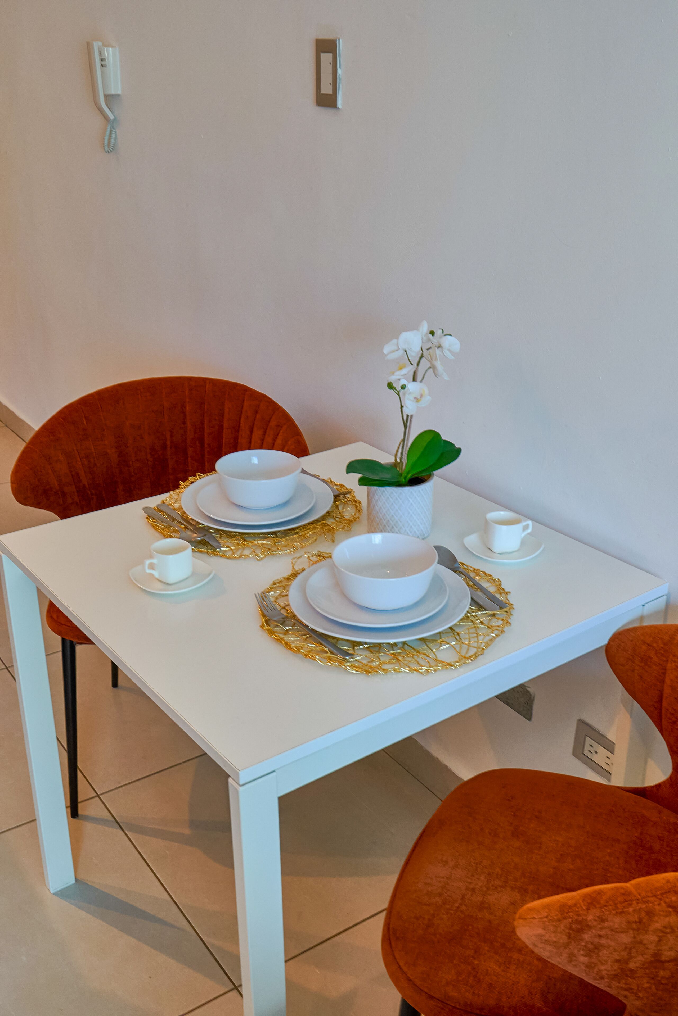 A dining table set with white plates, bowls, and cutlery, accompanied by a white cup and saucer, a gold placemat, and a potted orchid.