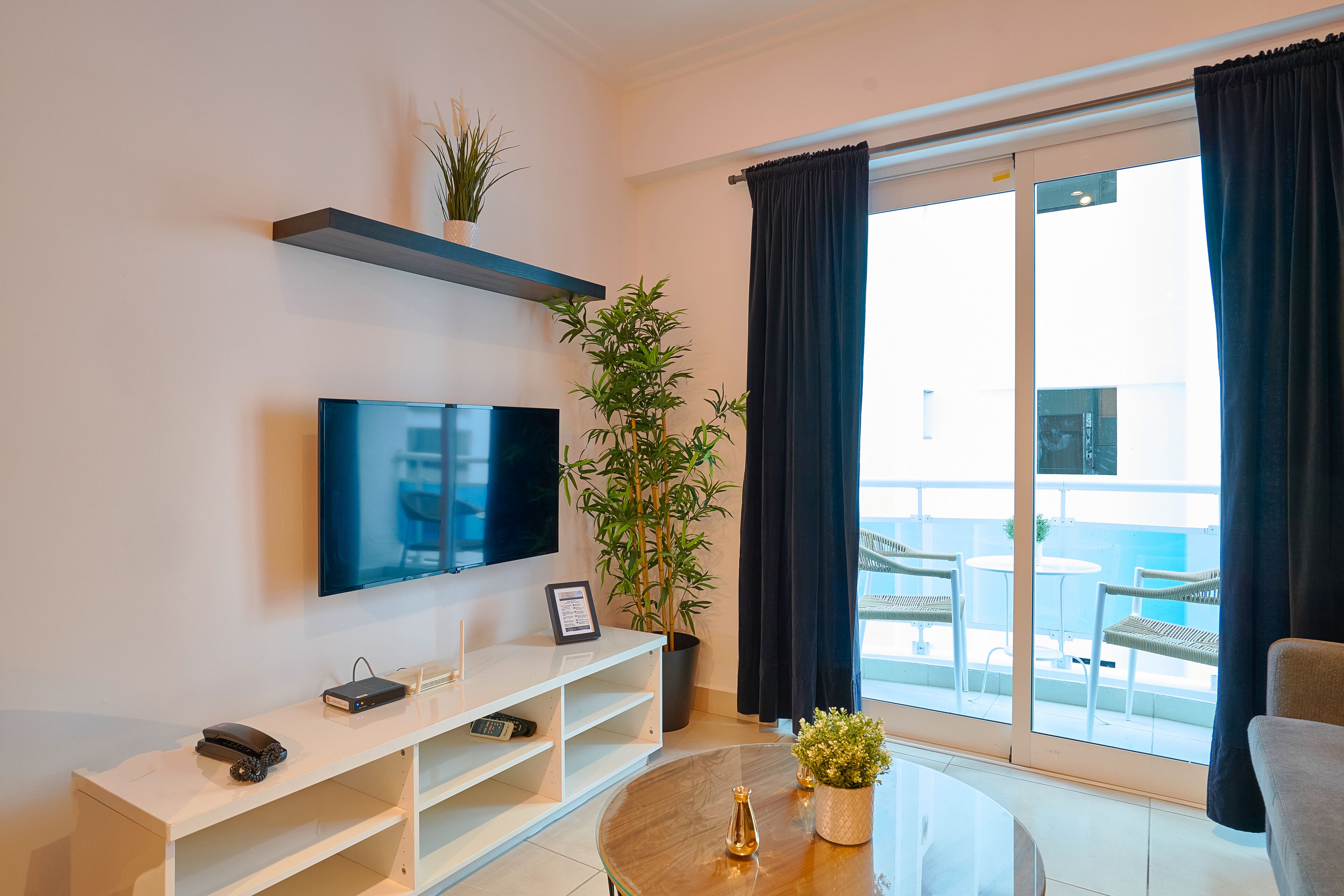 Uma sala de estar moderna com televisão de tela plana, um rack branco, uma planta em vaso e uma varanda com mobiliário de exterior.