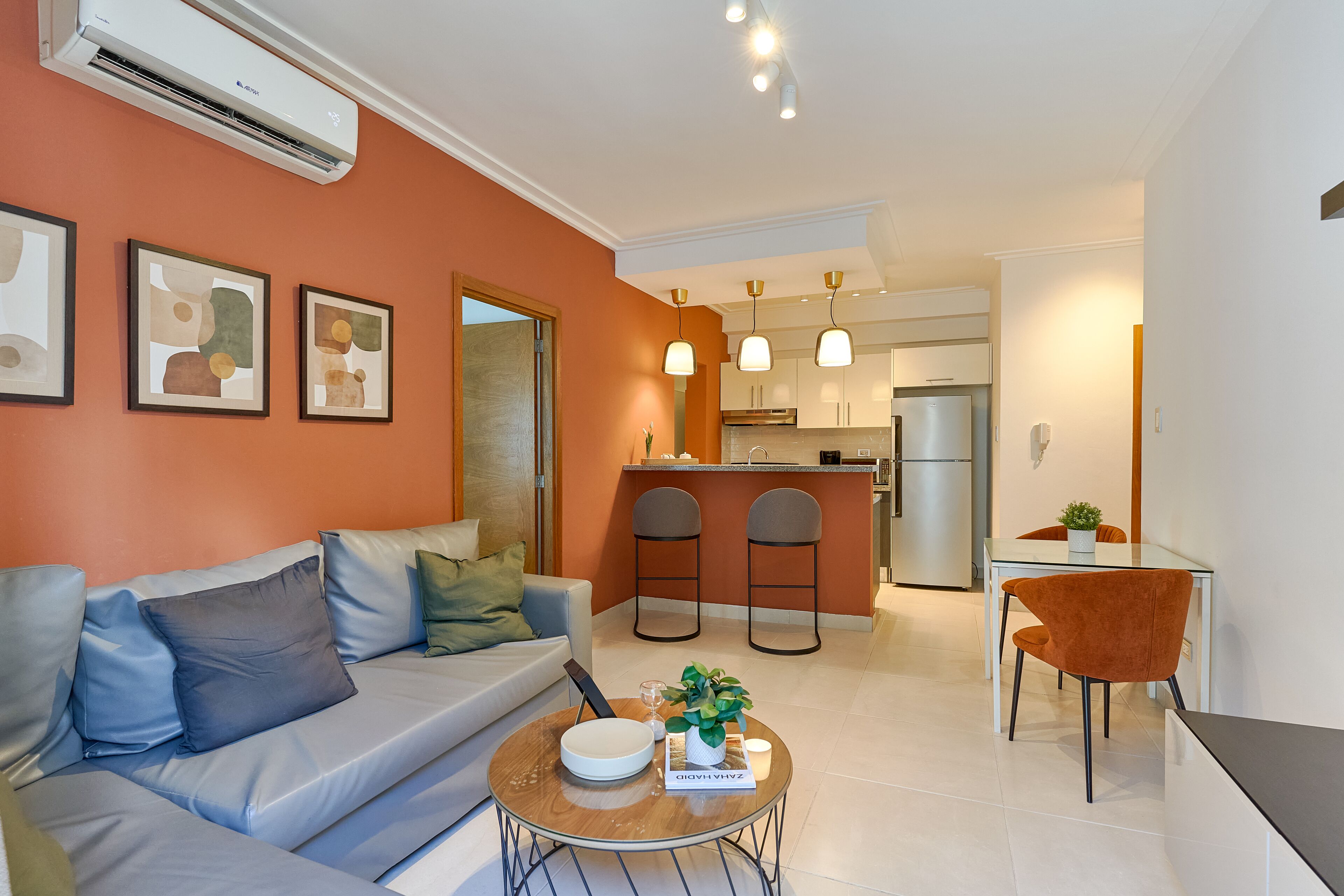 A modern living room with a sofa, a coffee table, and a dining area with a table and chairs. There is a kitchen in the background with a refrigerator and a bar counter.