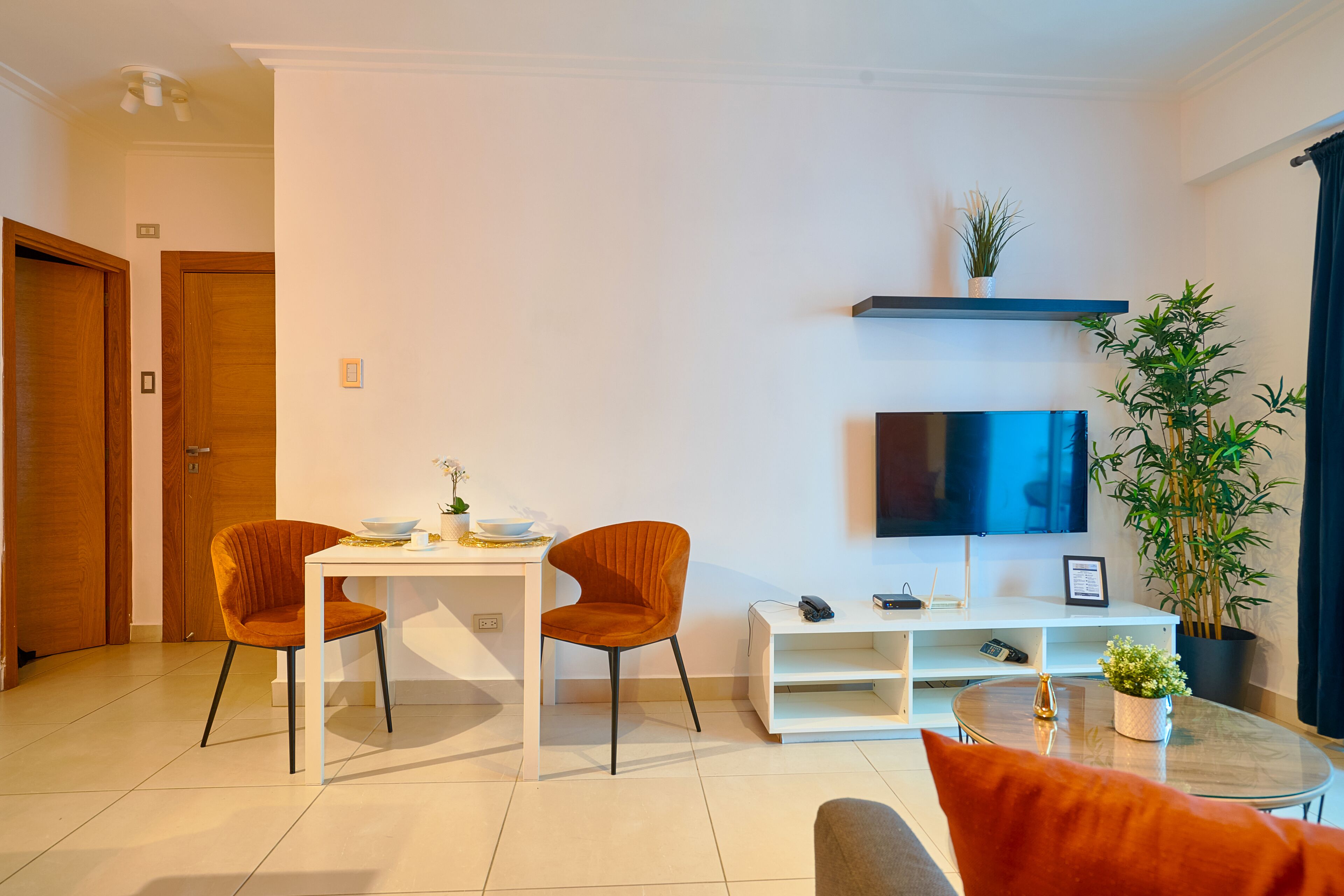 Una sala de estar moderna con un sofá blanco, dos sillas naranjas, una mesita, un televisor en la pared y una planta en maceta.