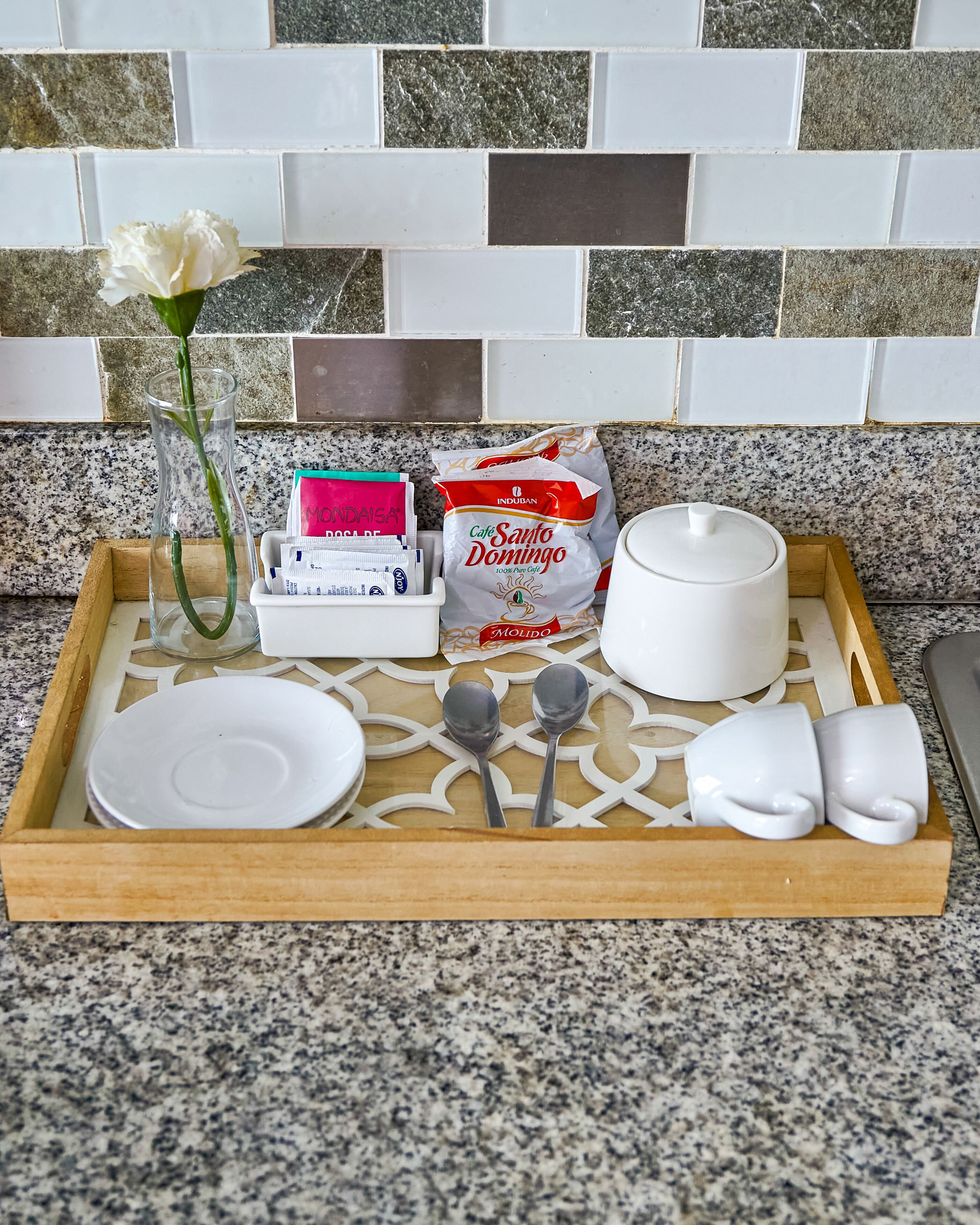 A tray with a white plate, two spoons, a white candle, a glass vase with a white flower, and a bag of 'Cafe Santo Domingo' coffee.