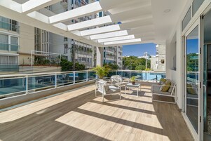 Un balcón moderno con suelo de madera, mobiliario blanco y barandilla de vidrio.