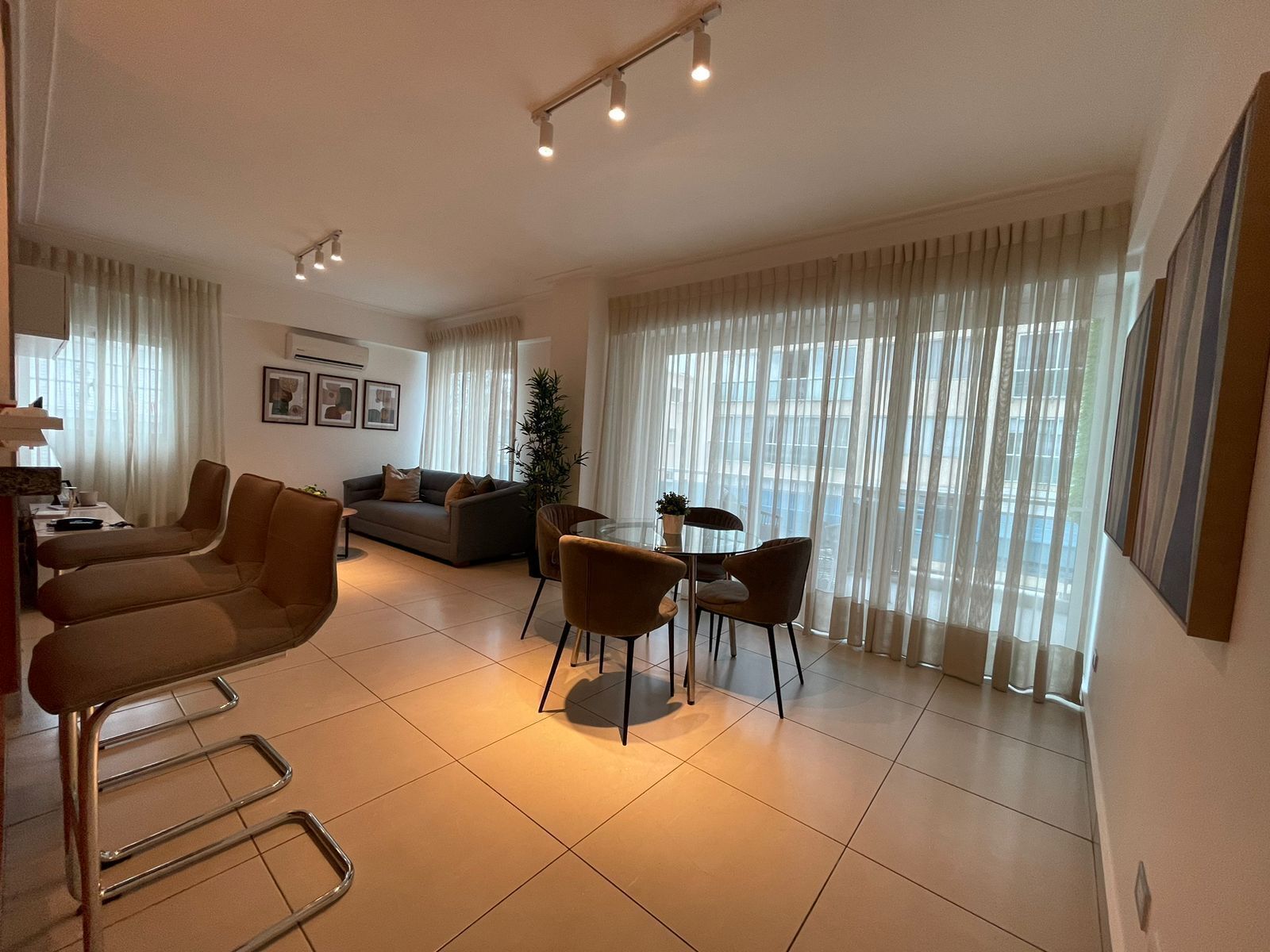 Un salon moderne comprenant un canapé, deux fauteuils, une table à manger avec des chaises et de grandes fenêtres ornées de rideaux légers.
