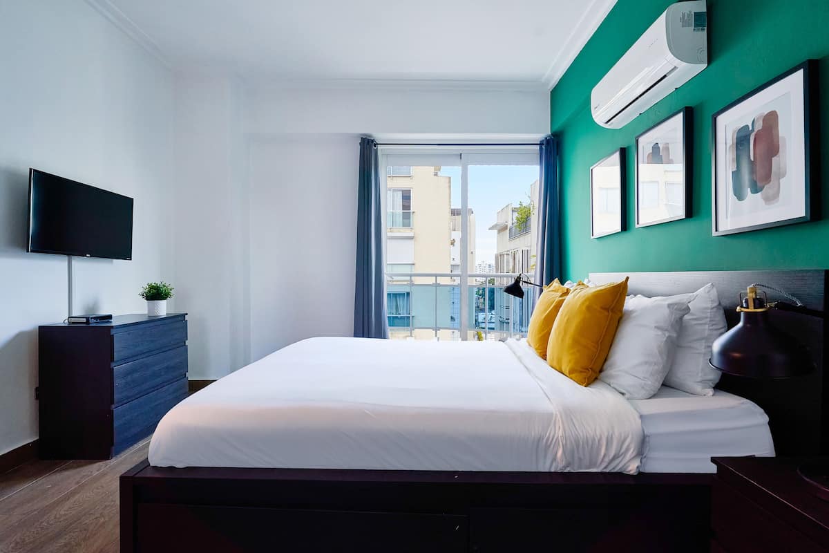 Een moderne slaapkamer met een groot bed, een flatscreen-tv, een dressoir en uitzicht op de stad door een groot raam.