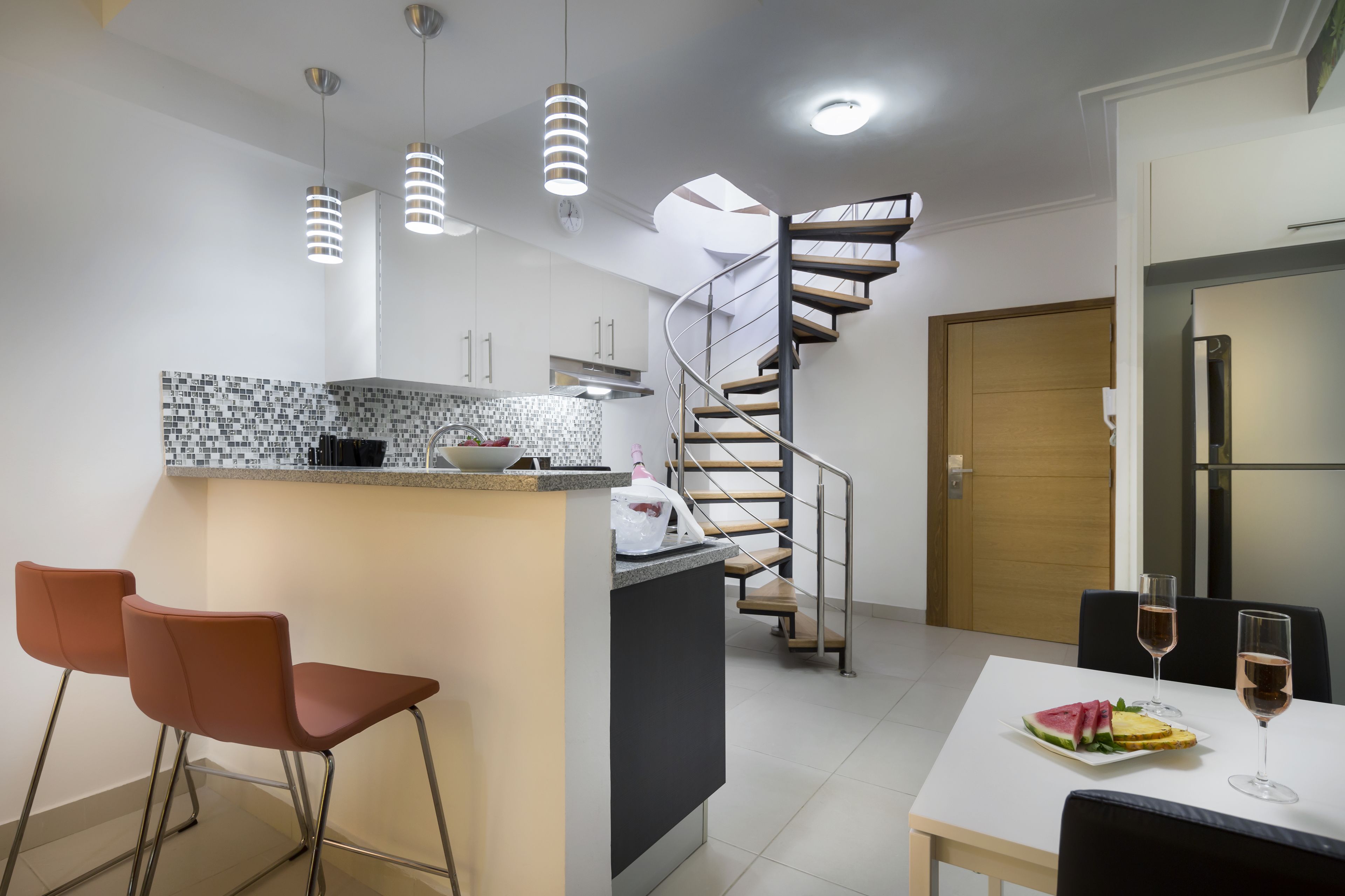 Une cuisine moderne dotée d’un îlot central, de deux tabourets de bar, d’une table à manger avec deux chaises et d’un escalier en colimaçon menant à un autre niveau.