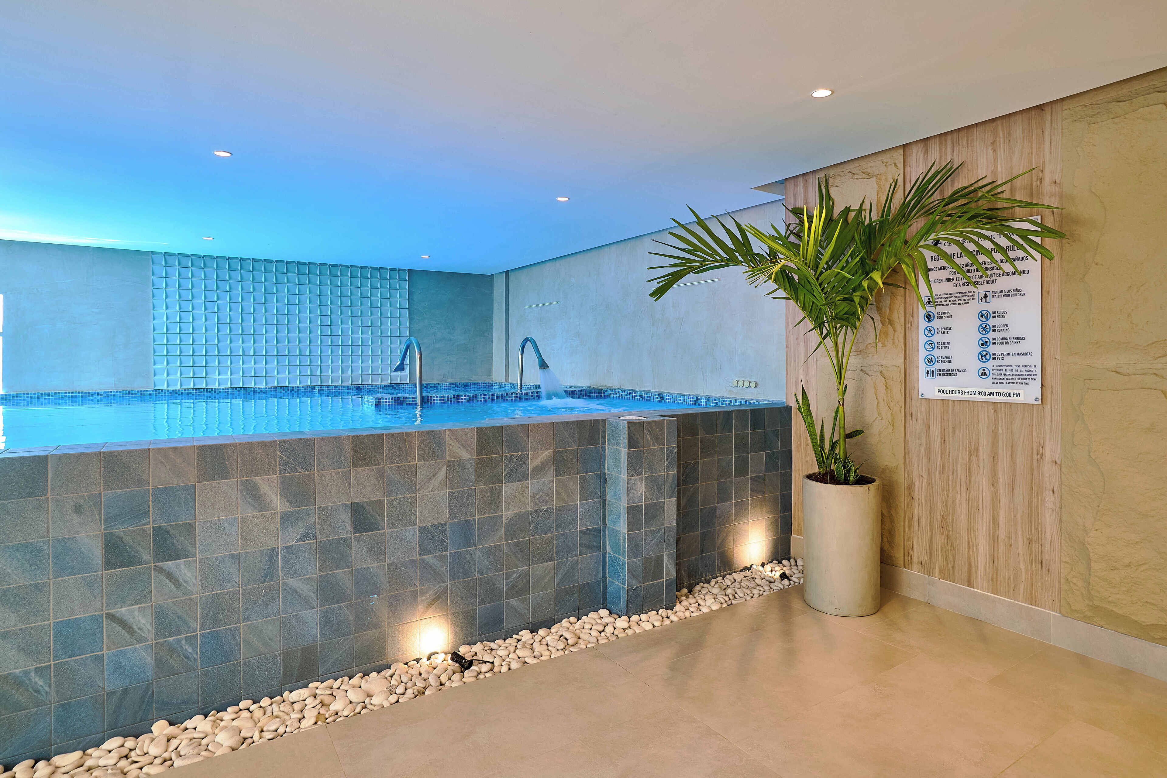 Un área de piscina interior moderna con un muro de azulejos, dos fuentes de agua y una planta en maceta.