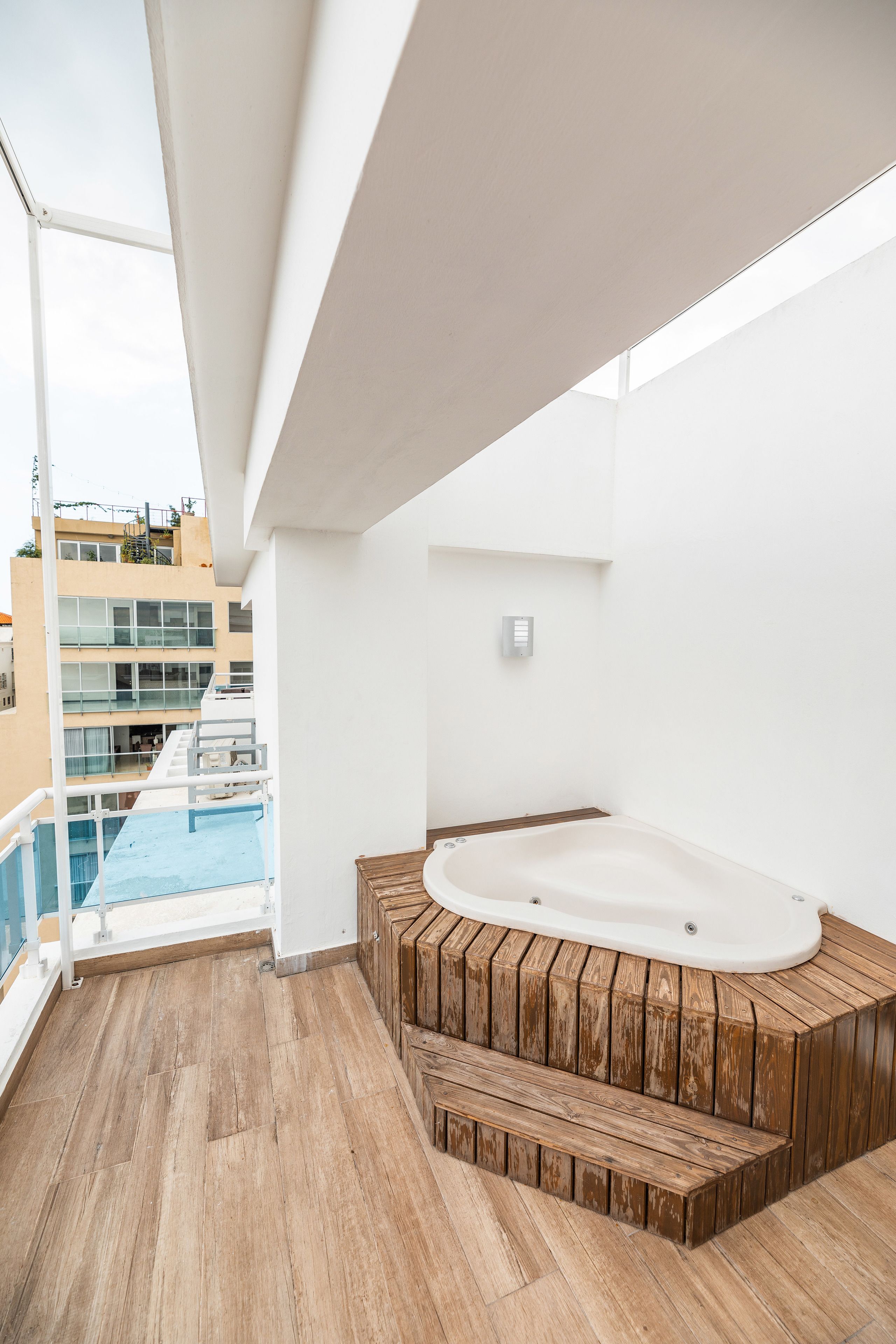 Un balcon moderne avec une terrasse en bois, un jacuzzi et une vue sur un immeuble à plusieurs étages doté de balcons.