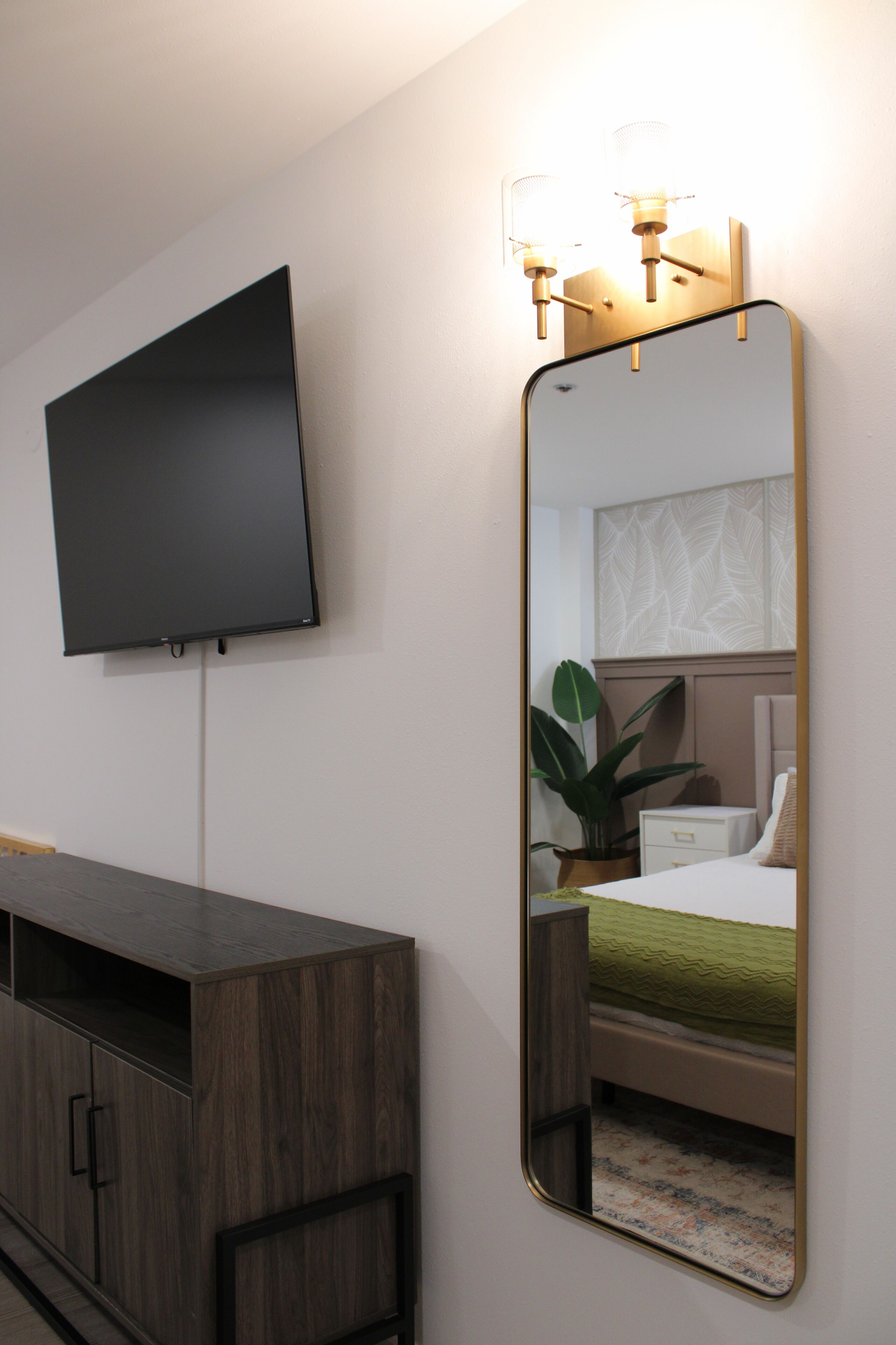 Ein modernes Hotelzimmer mit einem an der Wand befestigten Flachbildfernseher, einem Holzschrank, einem Nachttisch mit Pflanze und einem Bett mit grüner Bettdecke.