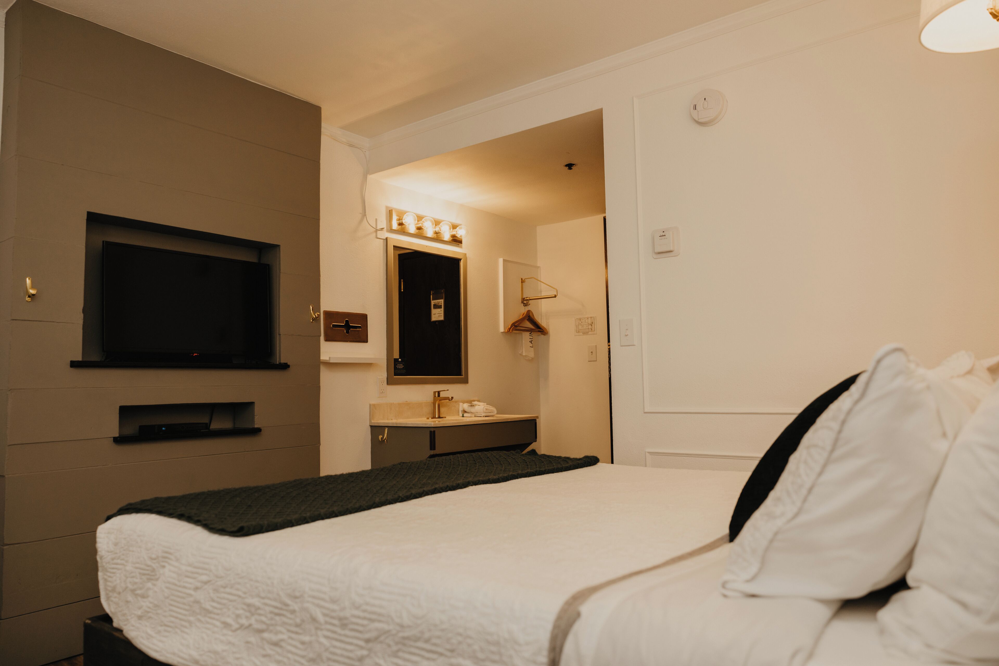 Habitación de hotel con cama, televisor montado en la pared, baño con espejo y lavamanos, y una puerta que da a otra habitación.