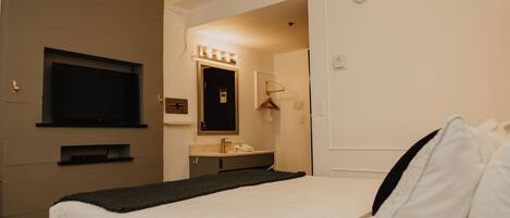 Habitación de hotel con cama, televisor montado en la pared, baño con espejo y lavamanos, y una puerta que da a otra habitación.