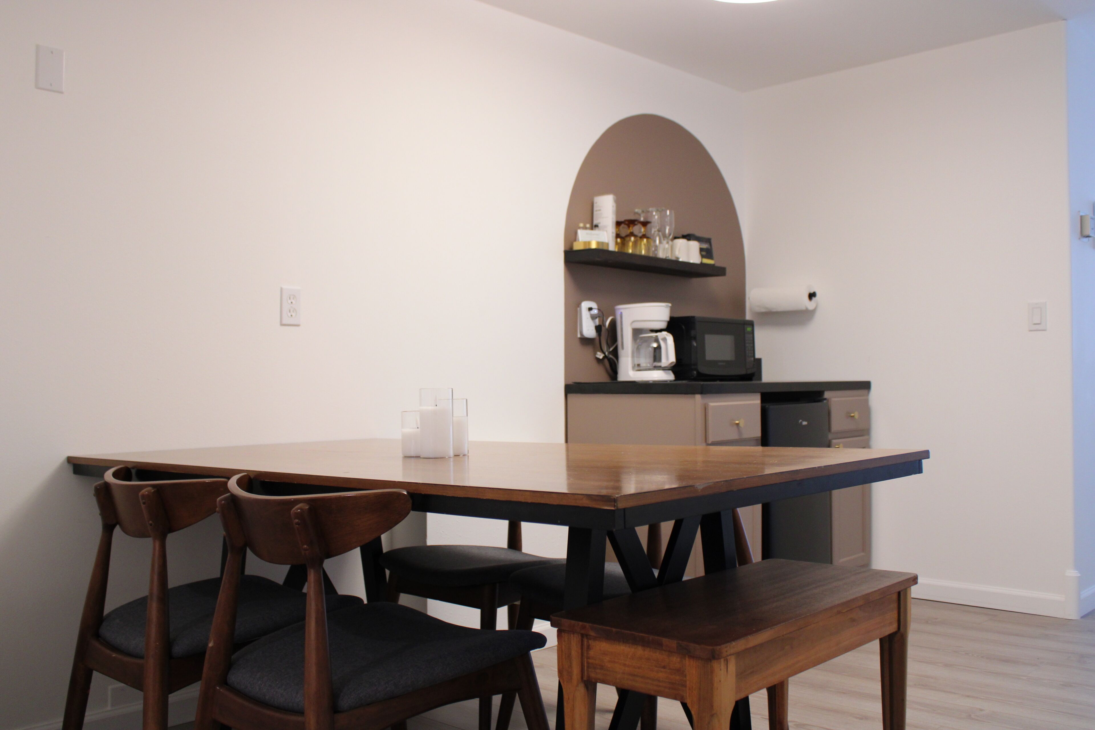 Un espace repas avec une table et des chaises en bois, un coin cuisine équipé d’appareils électroménagers, et un mur blanc agrémenté d’une porte en arc de cercle.