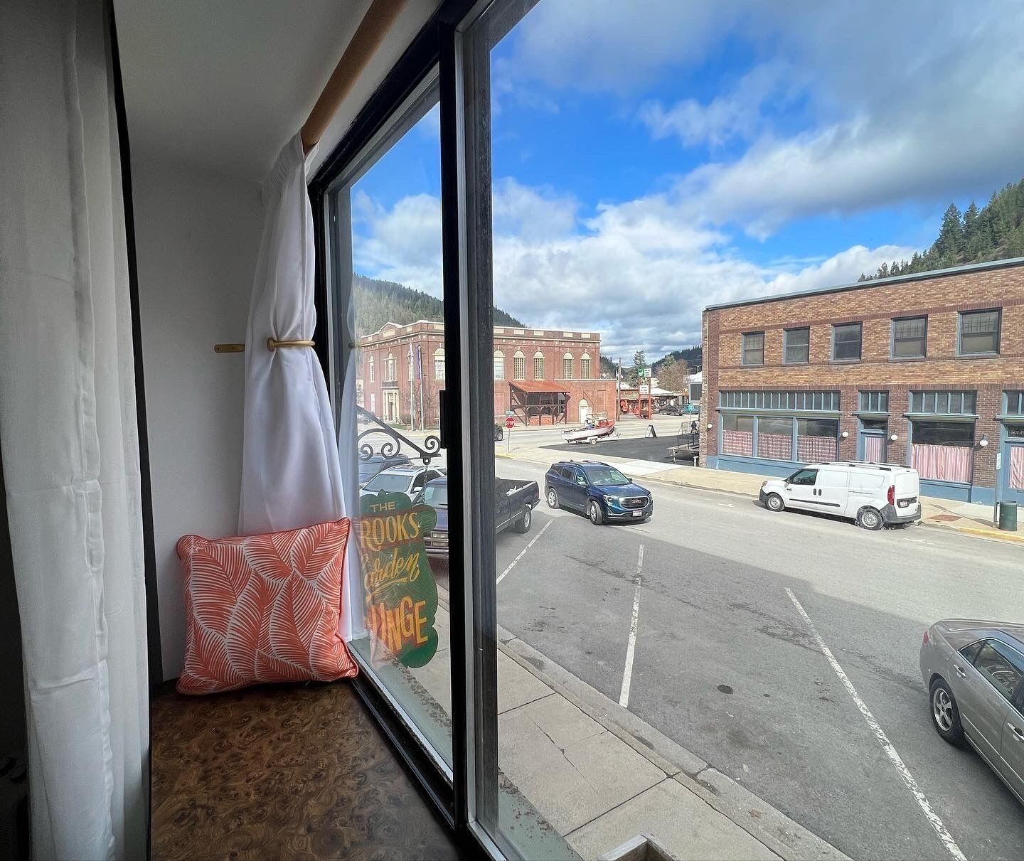 A view from a window with a pillow on a bench, a street with cars, and buildings in the background.