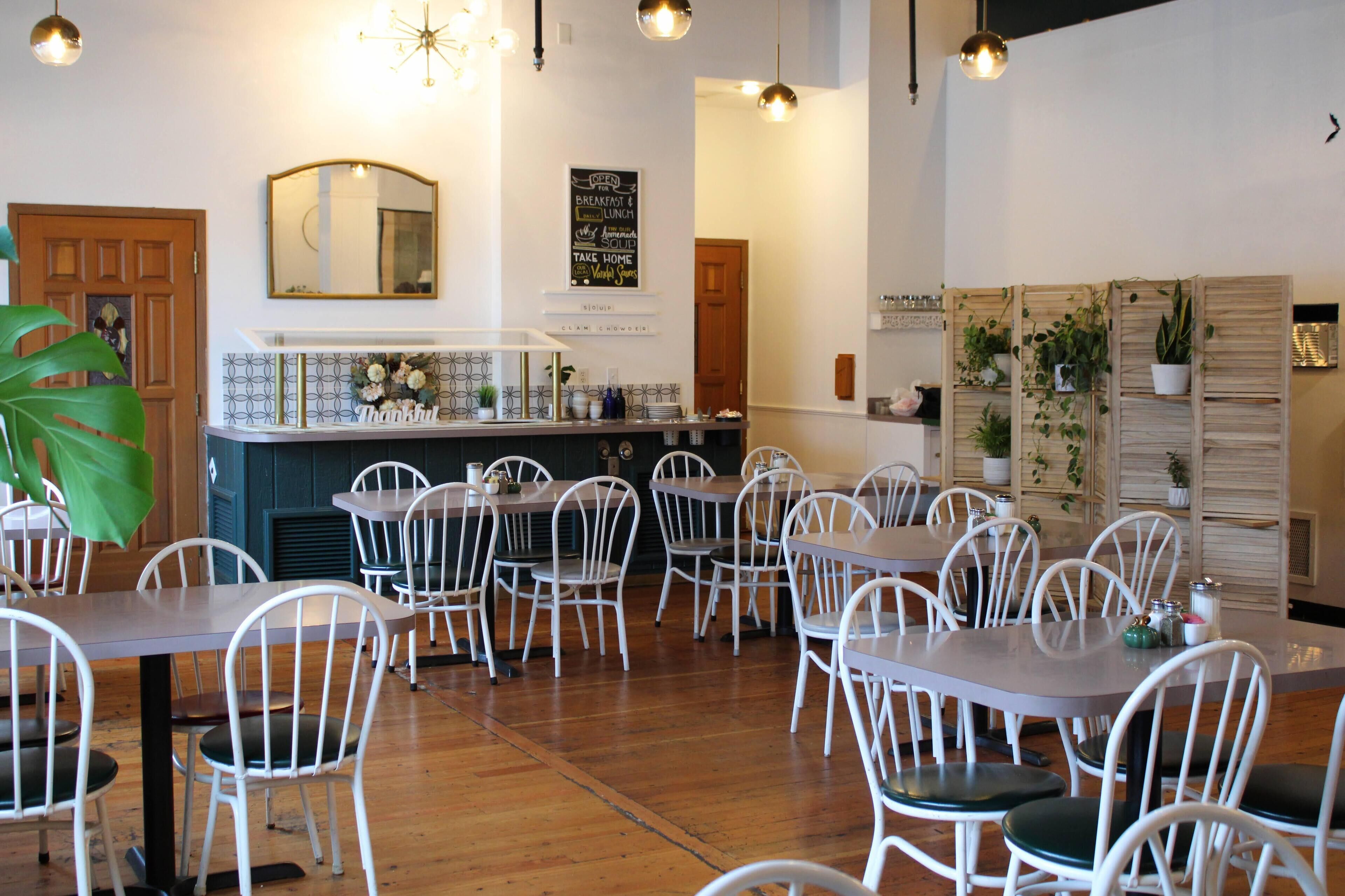 Un restaurante con sillas y mesas blancas, una barra con espejo y plantas en macetas sobre estantes.