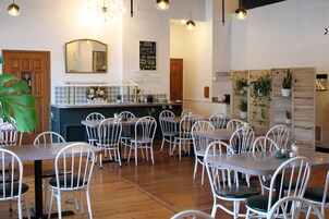 Un restaurante con sillas y mesas blancas, una barra con espejo y plantas en macetas sobre estantes.