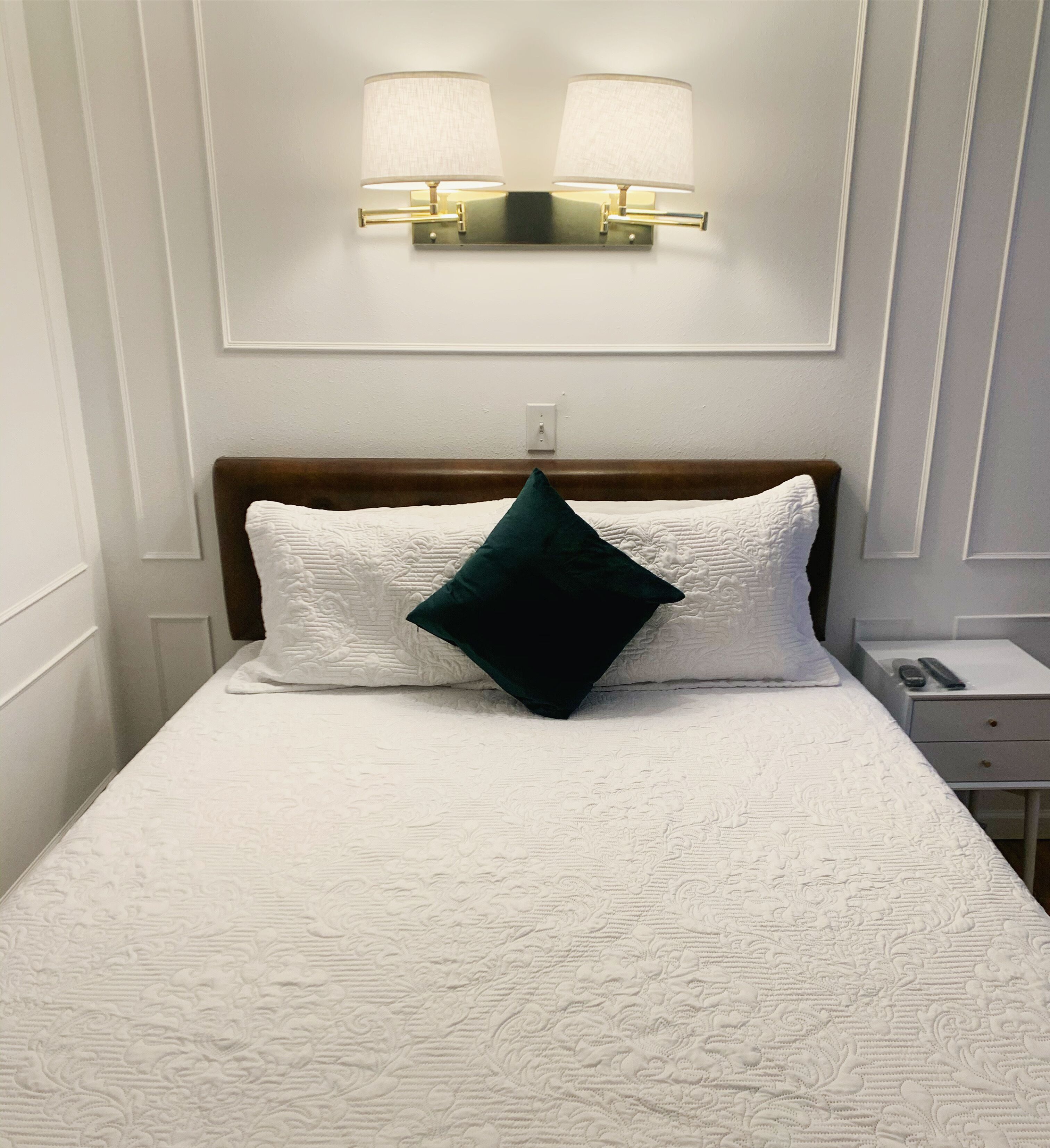 A neatly made bed with a white textured bedspread and a single dark green pillow. Above the bed, there is a wall-mounted light fixture with two lamps.