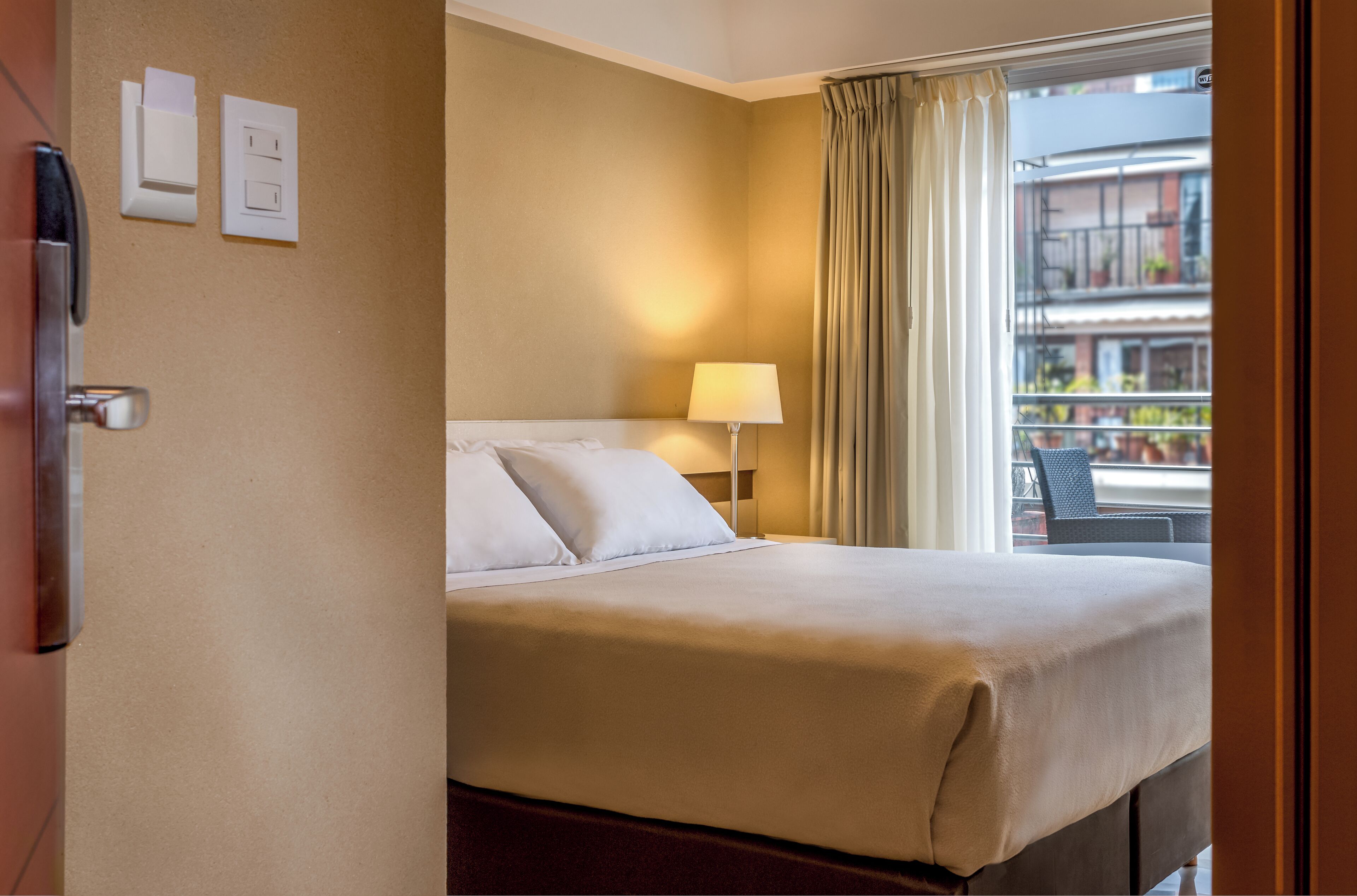 A hotel room with a bed, a lamp, a window with curtains, and a balcony view.