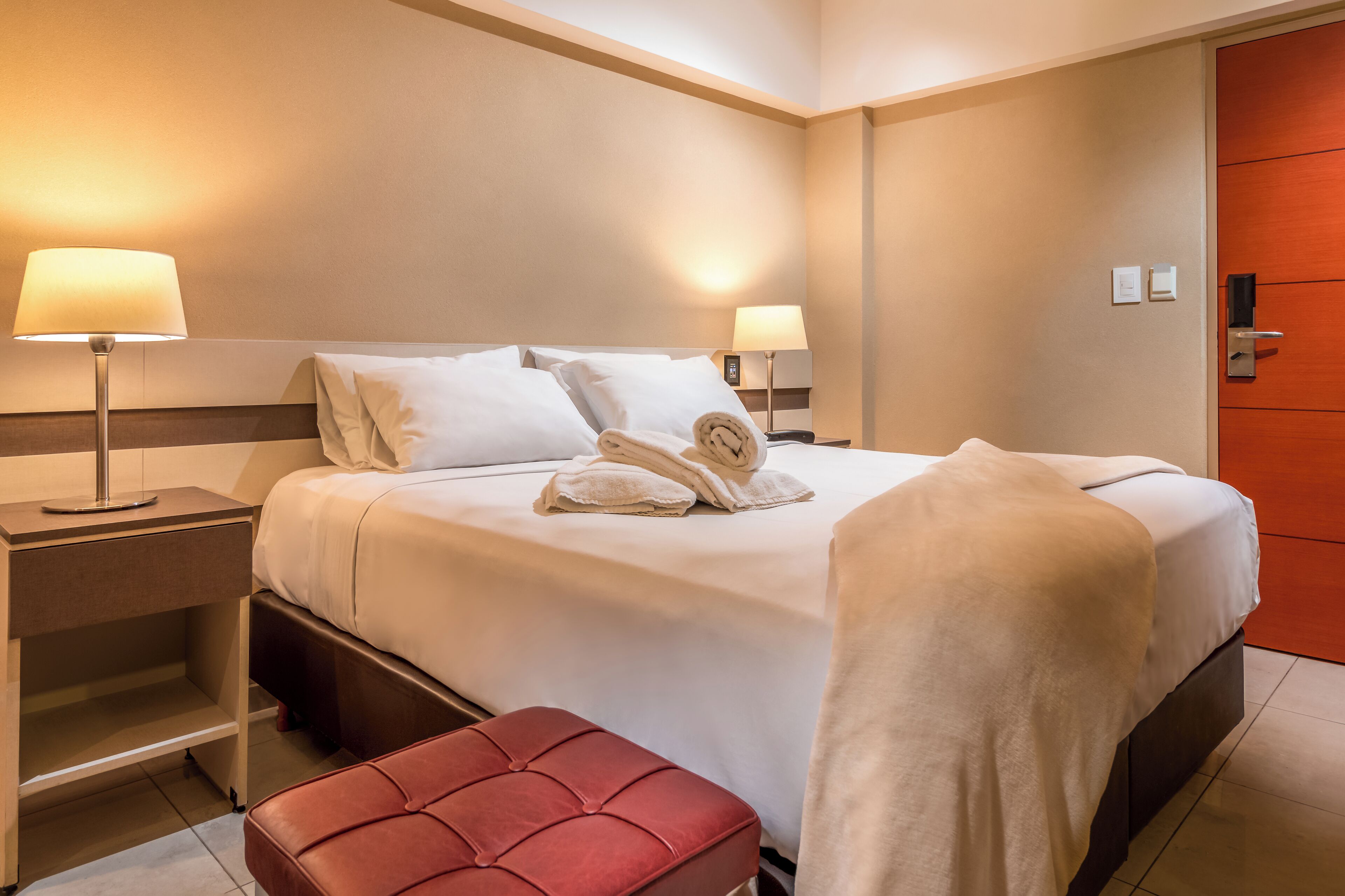 Habitación de hotel con una cama bien hecha, dos lámparas de noche, un banco rojo y una puerta con picaporte.