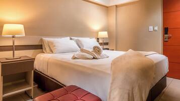 Habitación de hotel con una cama bien hecha, dos lámparas de noche, un banco rojo y una puerta con picaporte.