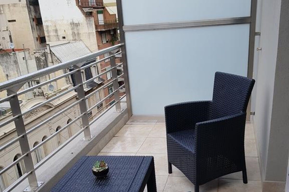 A balcony with two wicker chairs and a small table, overlooking a cityscape with buildings and a staircase.