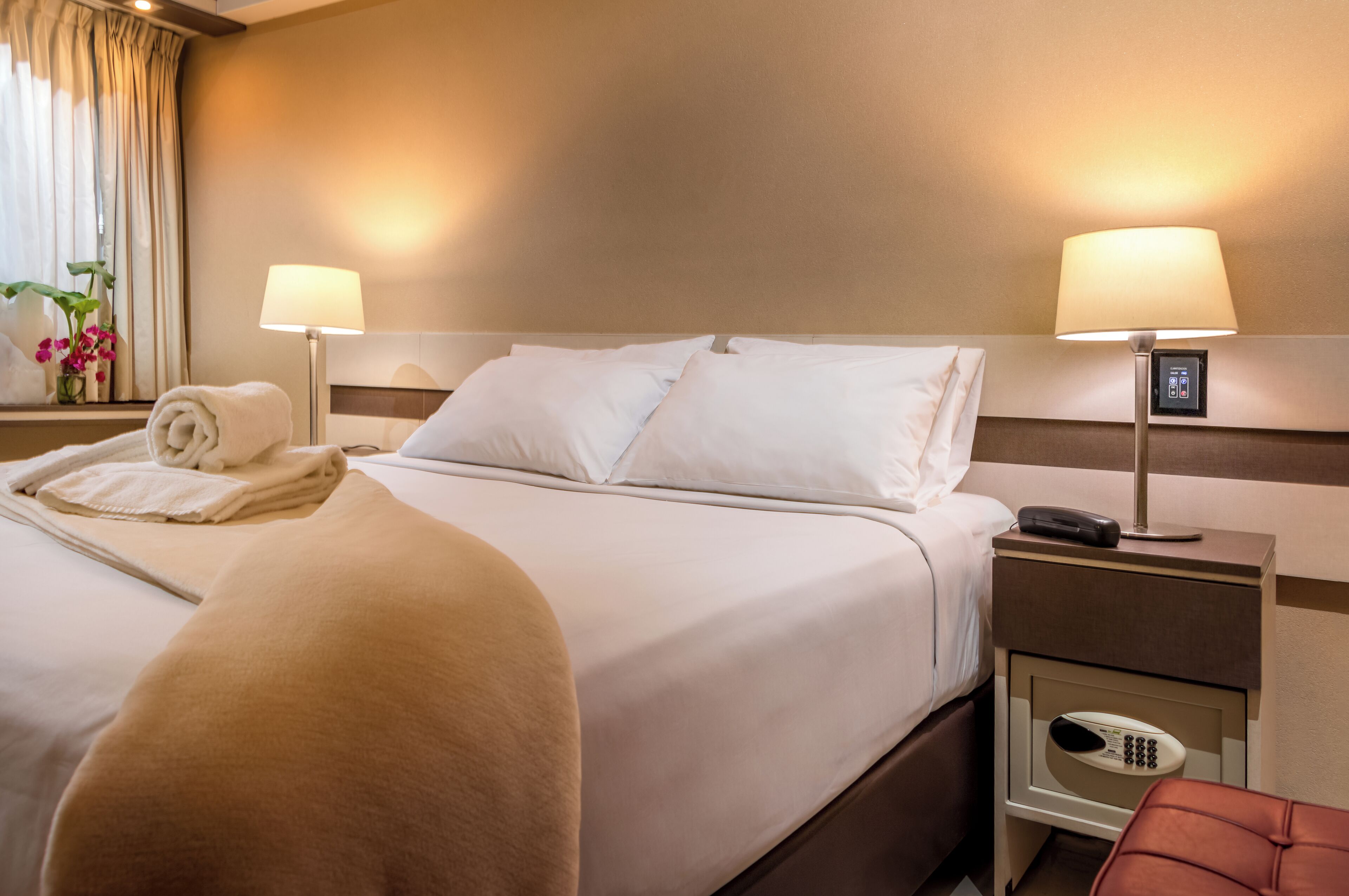 Habitación de hotel con una cama bien hecha, dos lámparas de noche, una mesita de noche con teléfono y una silla roja.