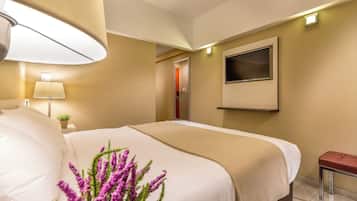Habitación de hotel con una cama, televisor montado en la pared, lámpara de noche y una mesita con un cojín rojo.