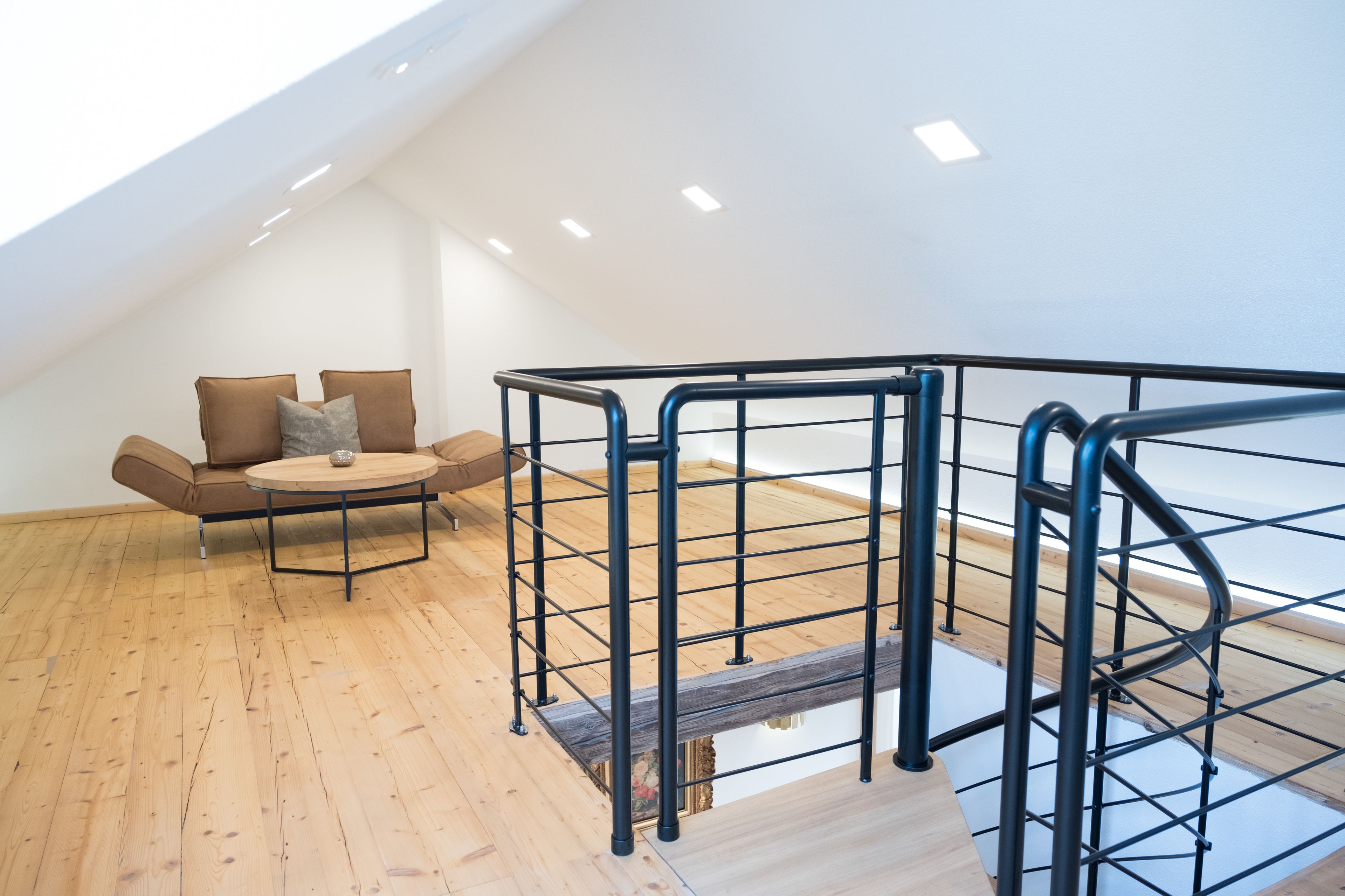 A modern loft bedroom with a wooden floor, a round wooden table, a sofa with cushions, and a metal staircase railing.