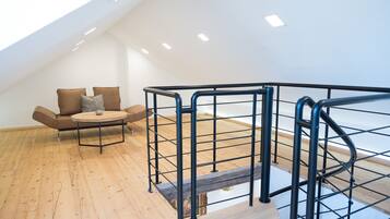 A modern loft bedroom with a wooden floor, a round wooden table, a sofa with cushions, and a metal staircase railing.