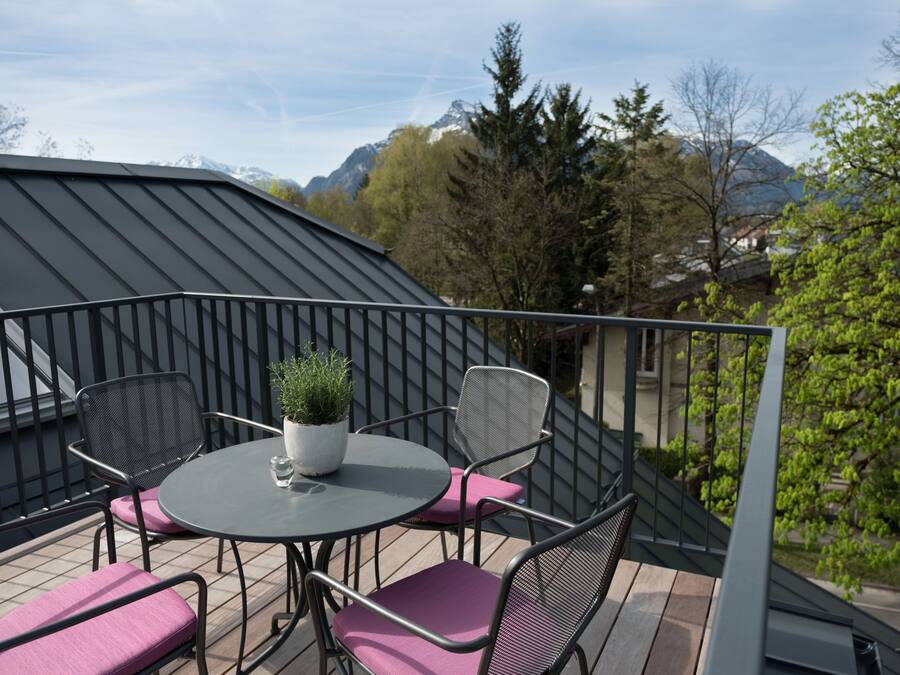 A balcony with a round table and chairs, a potted plant, and a view of mountains and trees.