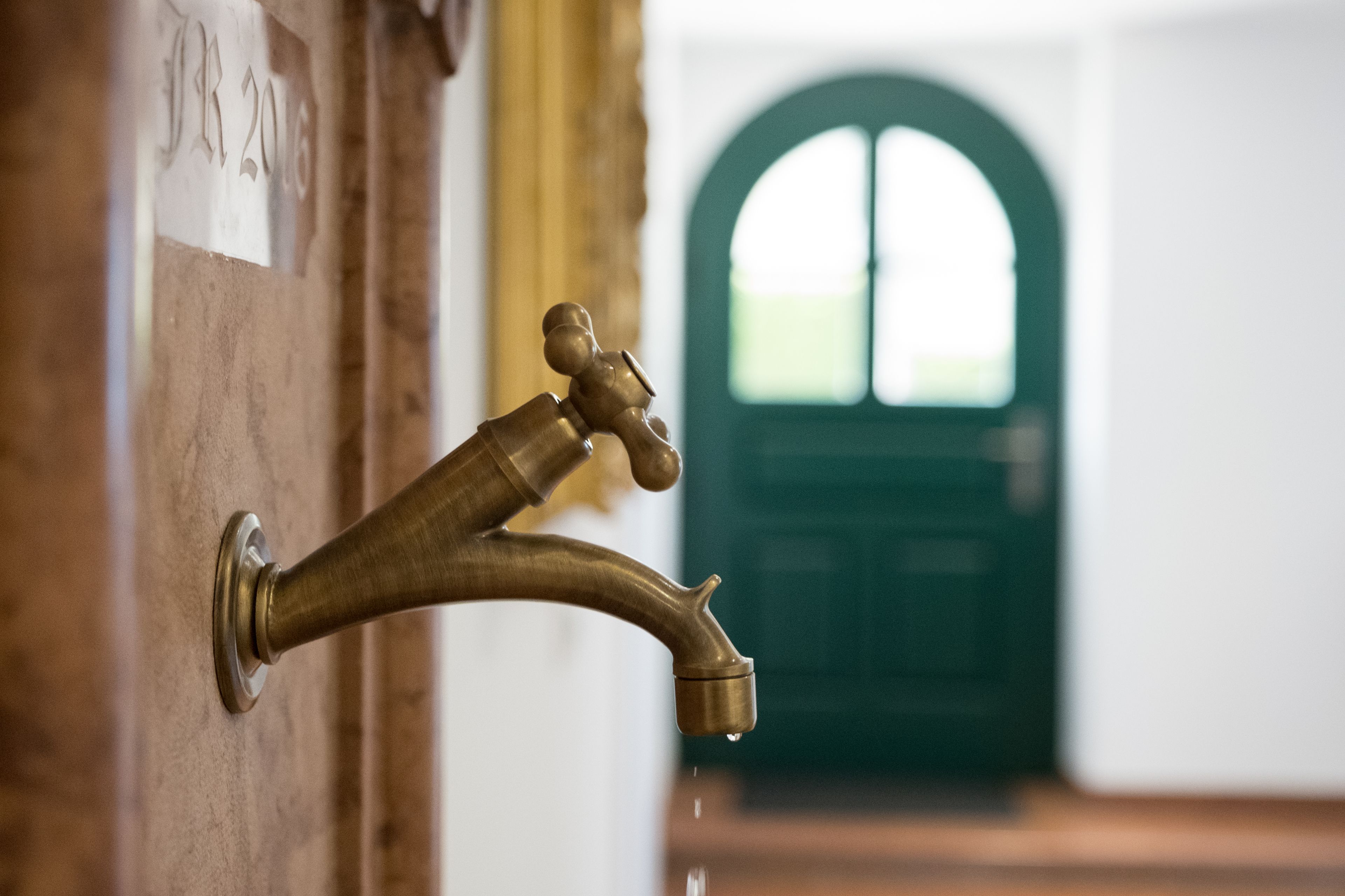 Un grifo de bronce con agua corriendo, fijado a una pared con una puerta al fondo.