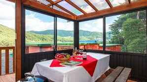 A table with a red tablecloth, a basket of fruit, and a bottle of wine on a balcony with large windows overlooking a mountainous landscape. - Castle of Magic Pension (Hoengseong)