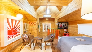 Quarto de cabana de madeira aconchegante, com uma cama, duas cadeiras de vime, uma mesa pequena, uma TV e uma janela com cortinas.