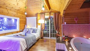 A cozy wooden cabin room with a bed, a small table with fruit, a bathtub, and a window with curtains. - Castle of Magic Pension (Hoengseong)
