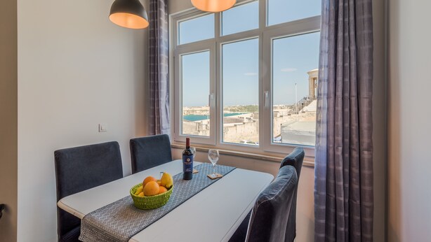 A dining area with a table set for two, a bowl of fruit, a bottle of wine, and a glass. The room has a large window with curtains, a view of the sea and buildings.