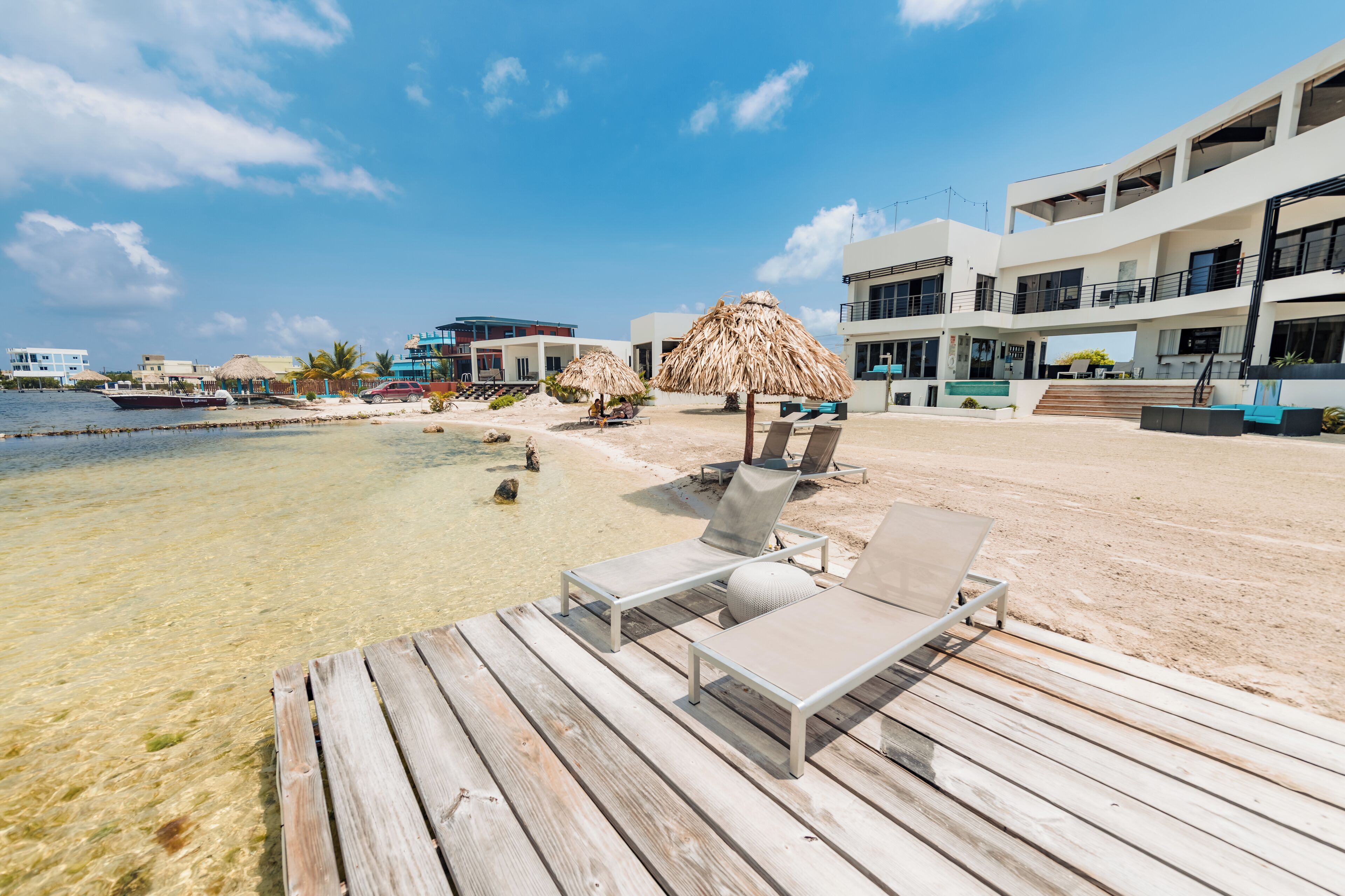 A wooden deck with lounge chairs and a thatched umbrella, overlooking a clear beach and a modern building with balconies.