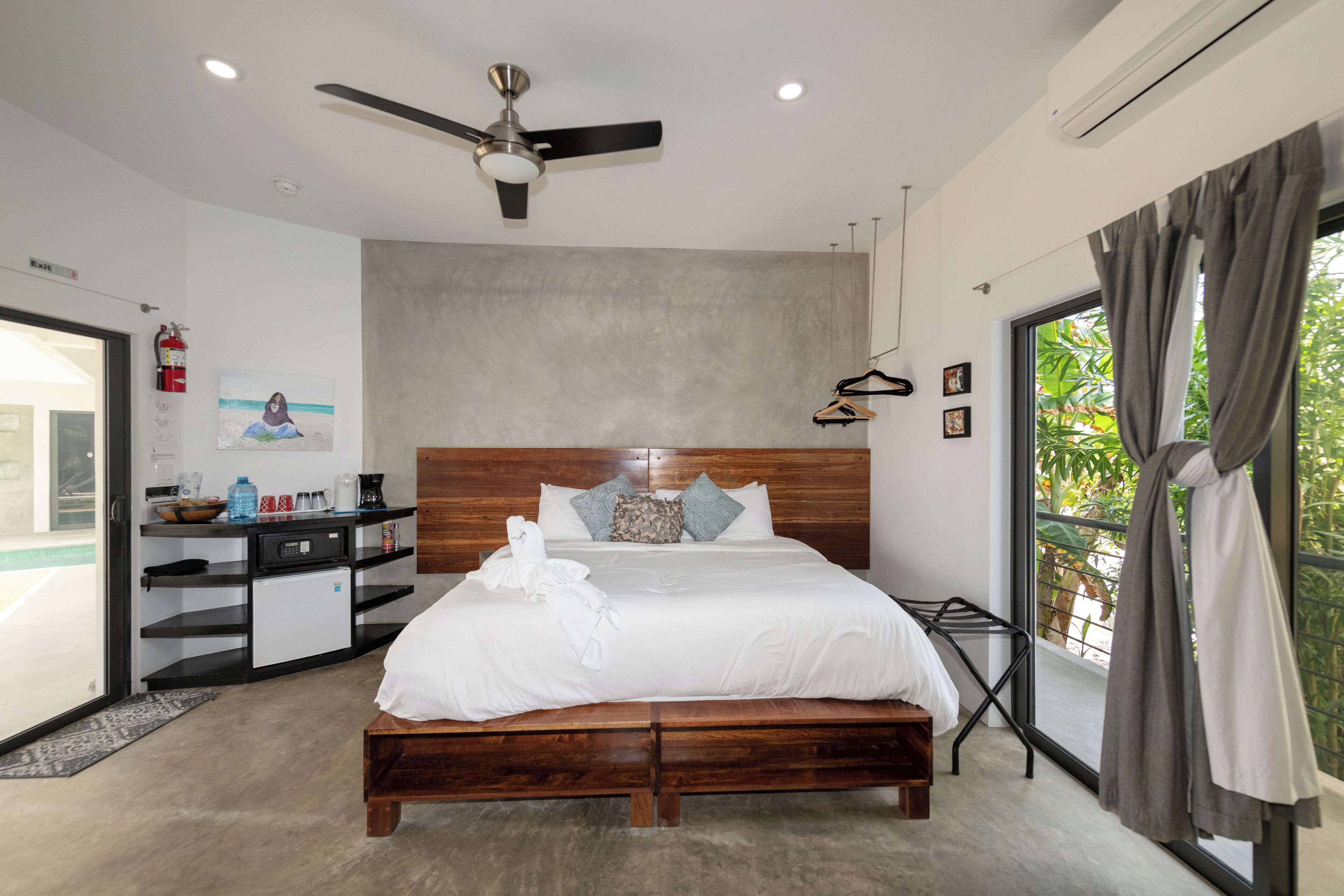 Un dormitorio moderno con cama de madera, ventilador de techo, kitchenette pequeño y balcón con cortinas.