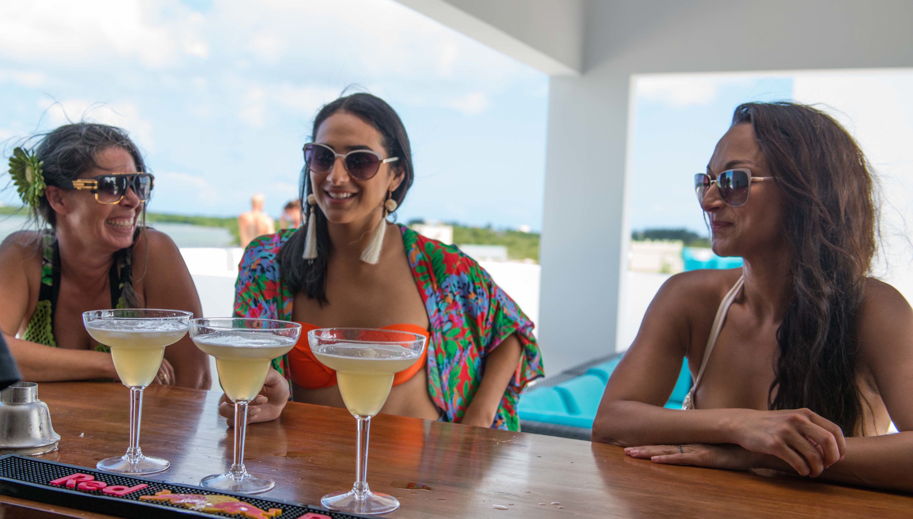 Three people sitting at a table with drinks, outdoors with a view of the beach.