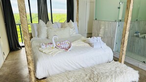 Uma cama com travesseiro e toalha em formato de cisne, um banquinho branco fofo e um banheiro com chuveiro de vidro.