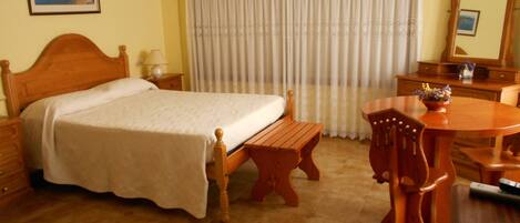 A hotel room with a wooden bed, a wooden bench, a round wooden table, and a chair. There is a mirror on the wall and a picture frame.