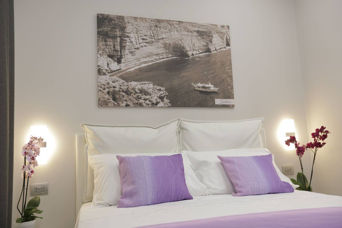 Una camera d'hotel con un letto, biancheria da letto bianca, cuscini viola, un quadro appeso al muro raffigurante un fiume e due orchidee in vaso.