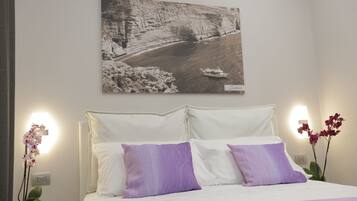 A hotel room with a bed, white bedding, purple pillows, a wall-mounted painting of a river, and two potted orchids.