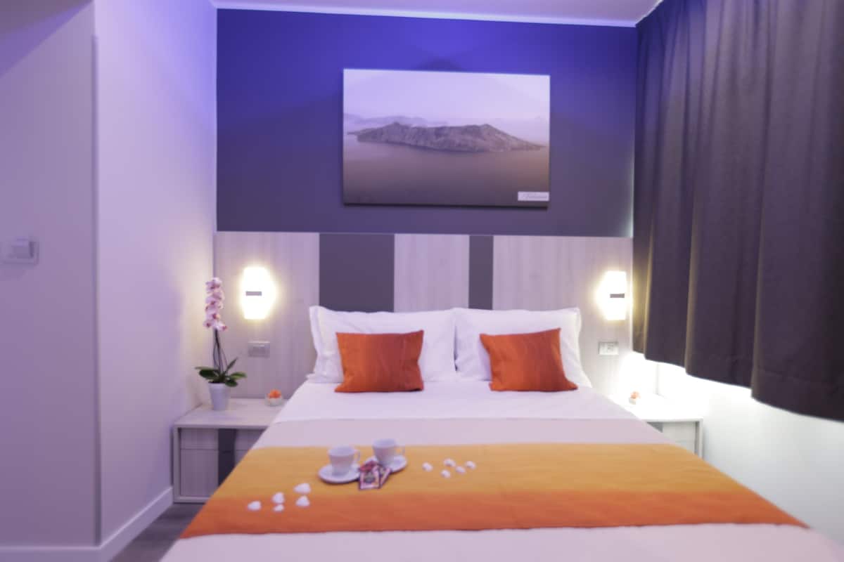 Una camera d'albergo con un letto, due comodini, un tavolino e una TV a parete, oltre a una finestra con tende.