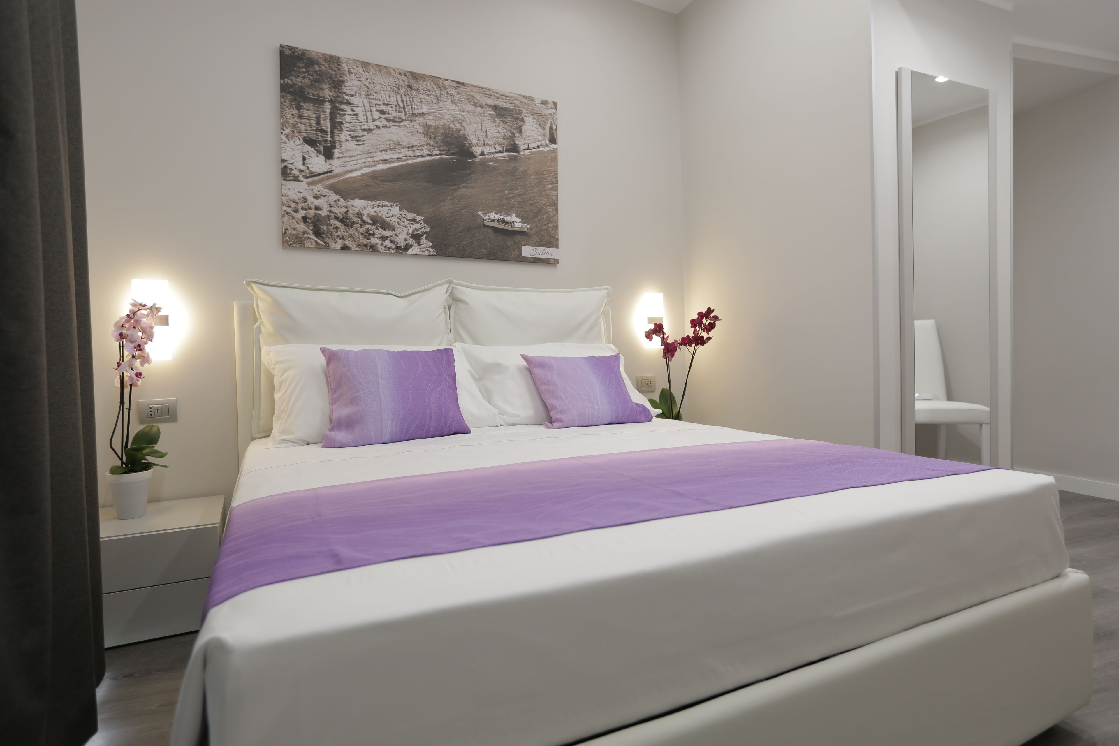 A modern bedroom with a large bed, white bedding, and purple accents. There is a painting on the wall and a small plant on the nightstand.