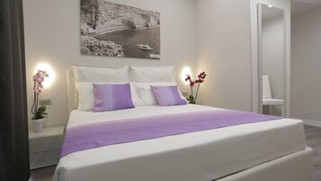 A modern bedroom with a large bed, white bedding, and purple accents. There is a painting on the wall and a small plant on the nightstand.