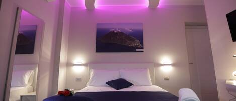 A bedroom with a bed, a nightstand, a mirror, a wall-mounted TV, and a painting of a mountain.