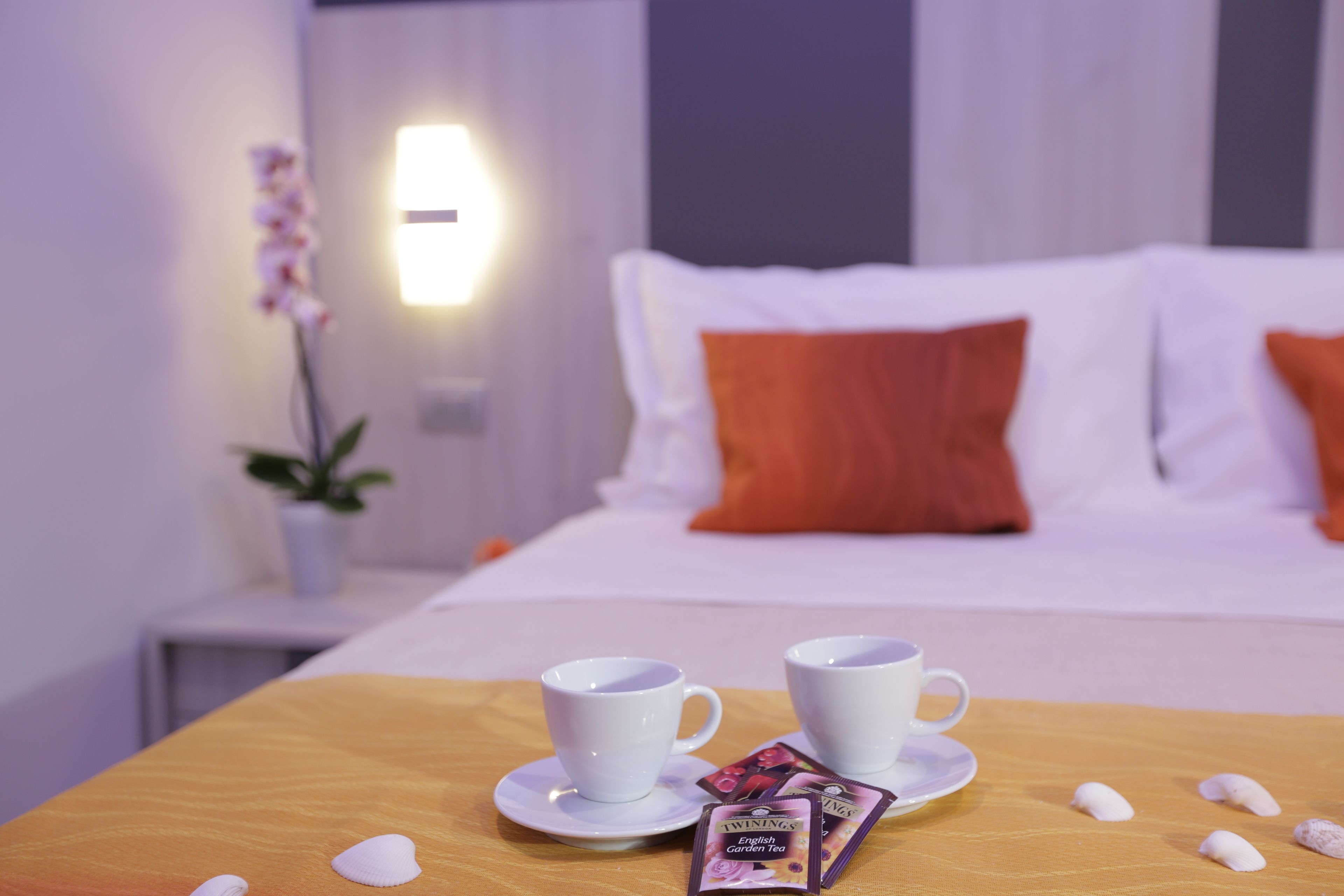 A neatly made bed with white and orange pillows, two white cups on a wooden table, and a packet of English Garden tea.